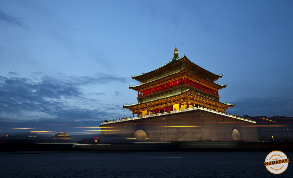 Vista nocturna del Campanario de Xi’an iluminado, con estelas de luces en movimiento al fondo y cielo azul profundo
