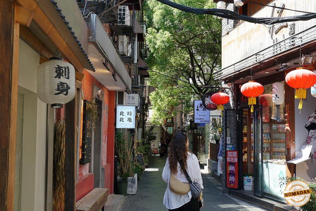 Tatiana caminando por un estrecho callejón en Shanghái, rodeado de fachadas tradicionales, tiendas locales y linternas rojas colgantes
