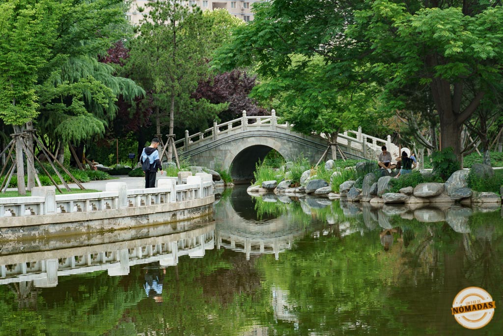 Puente de piedra arqueado reflejado en el agua en el Parque de la Muralla de Xi’an, rodeado de árboles y paseantes