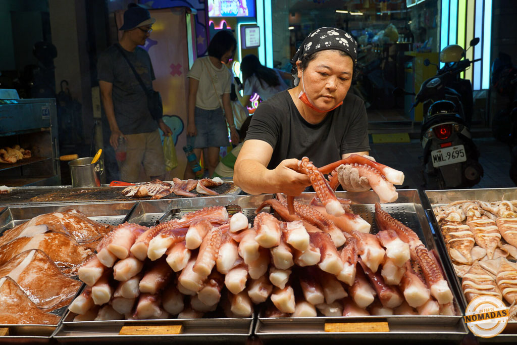 Vendedora taiwanesa preparando tentáculos de pulpo fresco en un mercado nocturno de Taipéi, con puestos callejeros al fondo.