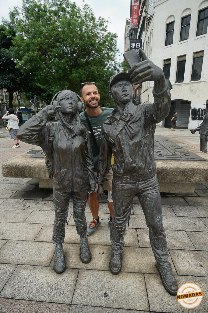 Oscar sonriente tomándose una foto junto a dos estatuas modernas en una calle de Taipéi.