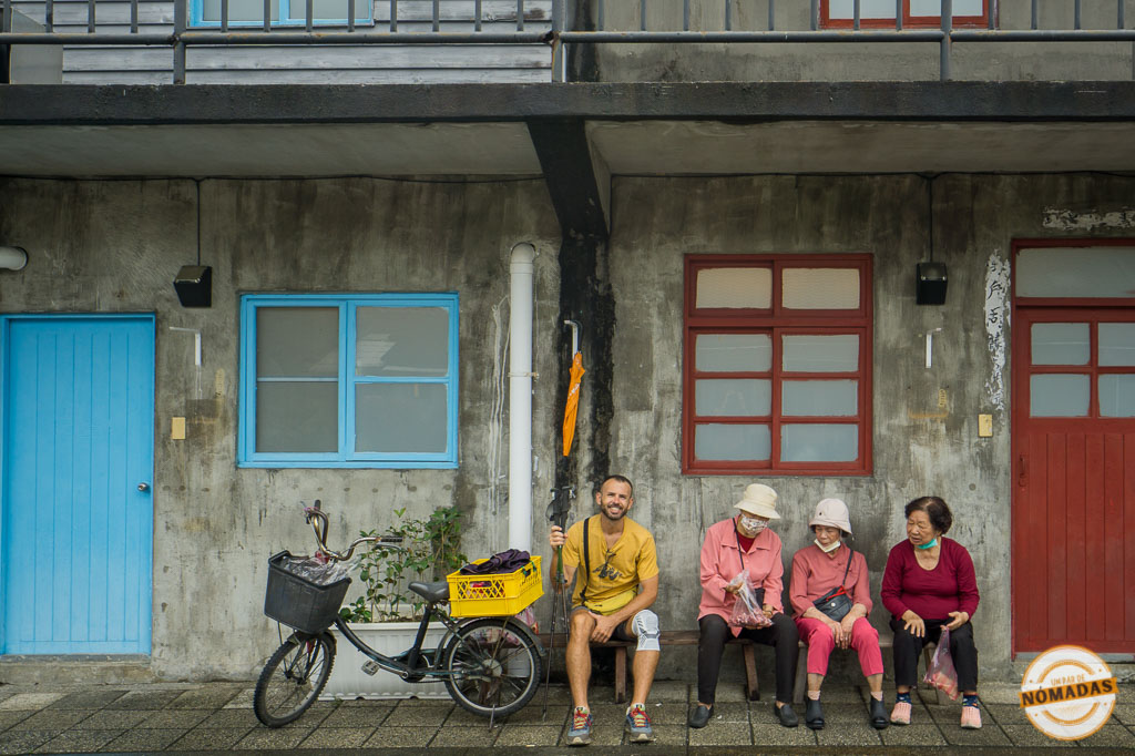 Escena de vida cotidiana en Taiwán con un viajero sentado junto a mujeres mayores locales en una calle de Taipéi, ideal para conocer la cultura auténtica del país