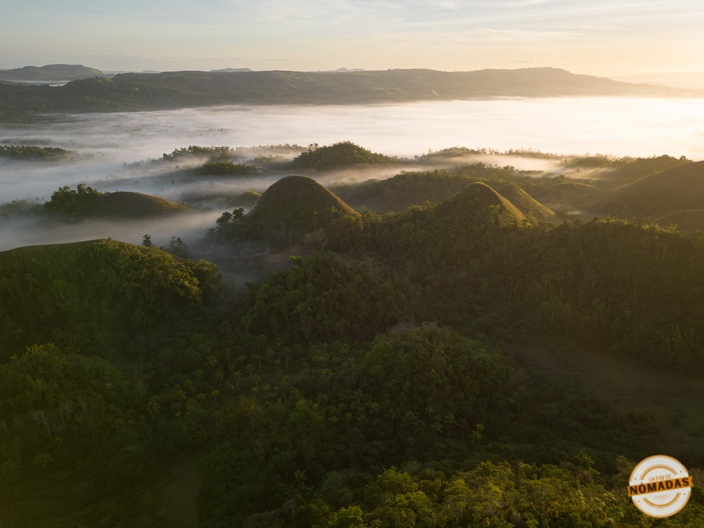 Fotografía aérea de las Chocolate Hills en Bohol, Filipinas, cubiertas de niebla al amanecer, con luz suave sobre las colinas cónicas.