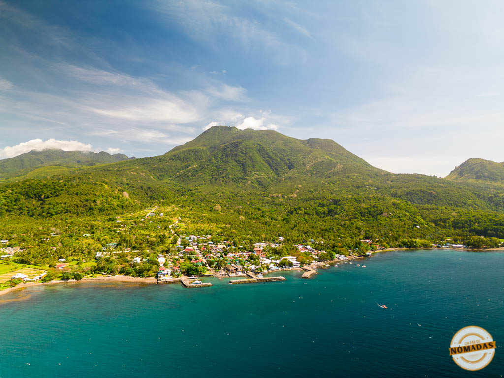 Imagen aérea de la costa de Camiguin, Filipinas, con un pequeño pueblo junto al mar y un volcán cubierto de vegetación en el fondo.