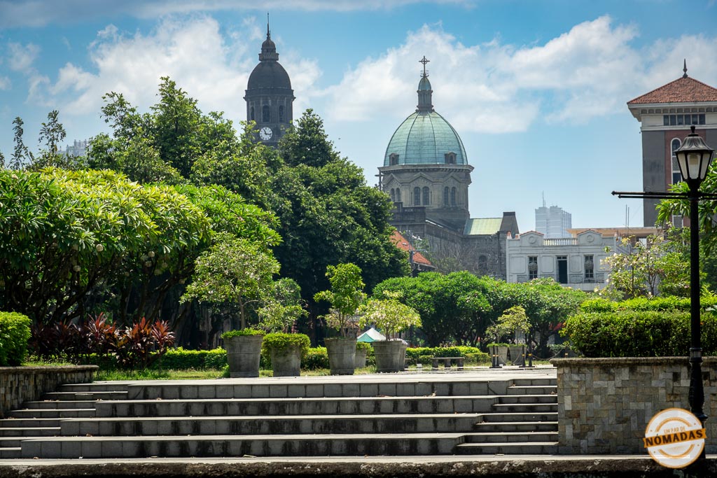 Vista de la Catedral de Manila desde los jardines de Intramuros, rodeada de vegetación y edificios coloniales.