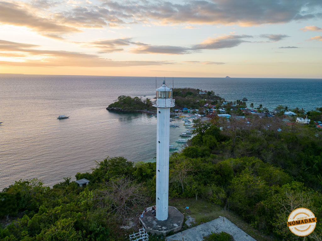 Vista aérea del faro blanco de Malapascua con el mar al fondo y el sol ocultándose, junto al pequeño pueblo costero de la isla.