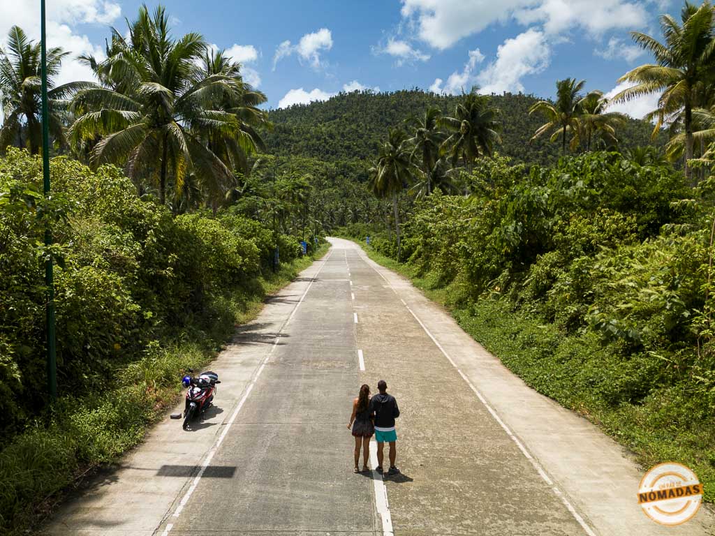 unpardenomadas caminando por una carretera rodeada de palmeras y vegetación tropical en Siargao, Filipinas, junto a su moto aparcada.