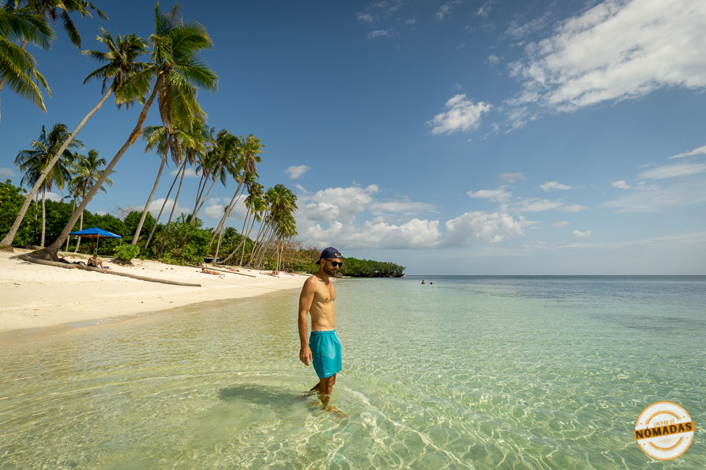 Oscar caminando por aguas cristalinas junto a una playa de palmeras en Siquijor, Filipinas, bajo cielo azul.