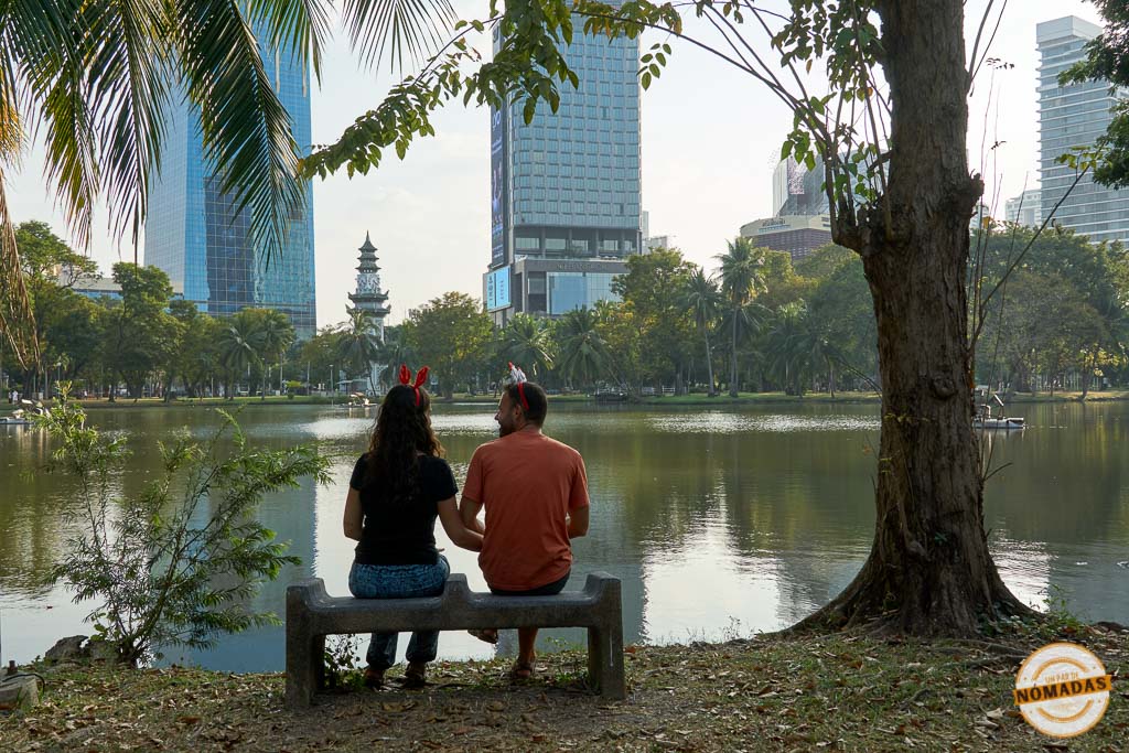 un par de nomadas sentados en banco frente al lago en Lumphini Park con edificios al fondo