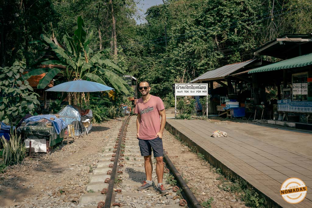 Oscar sobre las vías en la estación Nam Tok Sai Yok Noi, rodeada de vegetación y puestos locales en Tailandia
