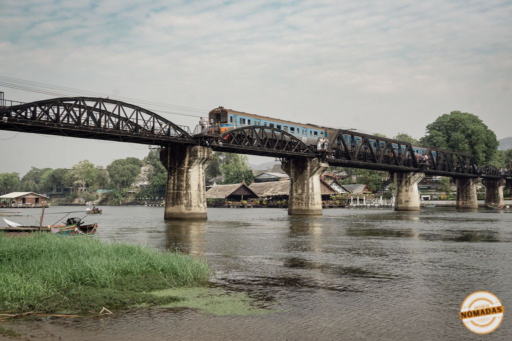 Tren azul cruzando el icónico puente sobre el río Kwai en Kanchanaburi, Tailandia, visto desde la orilla del río