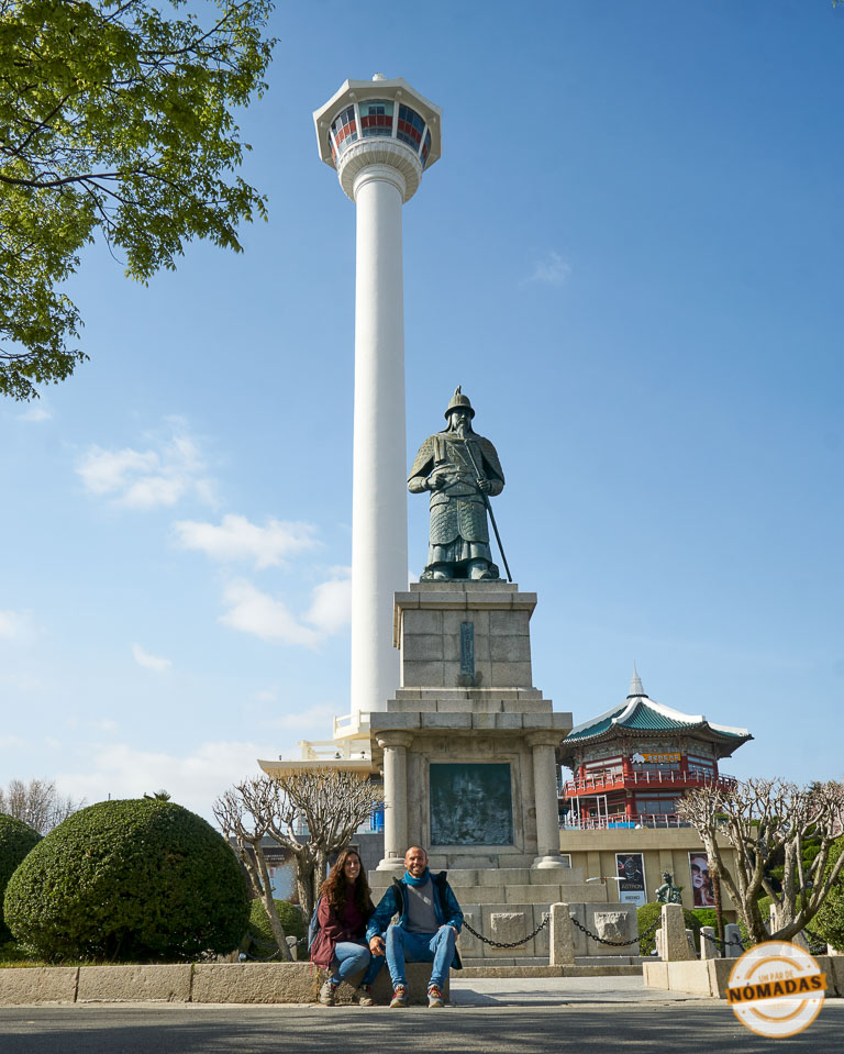 Pareja de viajeros frente a la estatua del almirante Yi Sun-sin y la Torre de Busán en Yongdusan Park, uno de los sitios más emblemáticos que ver en Busán
