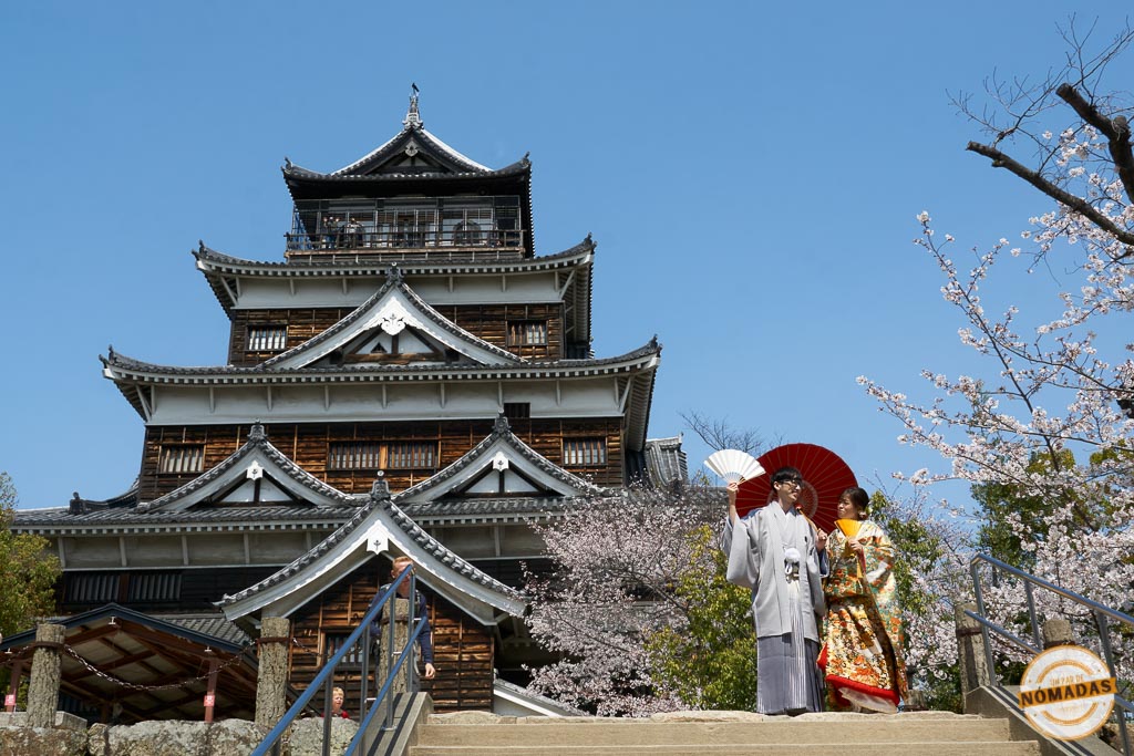 Pareja en kimono tradicional frente al castillo con sakura de fondo.