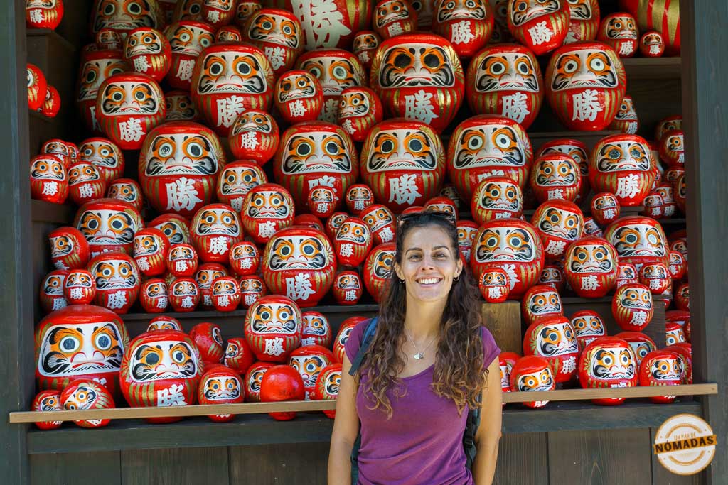 Tatiana sonriendo frente a estantería llena de muñecos Daruma rojos.
