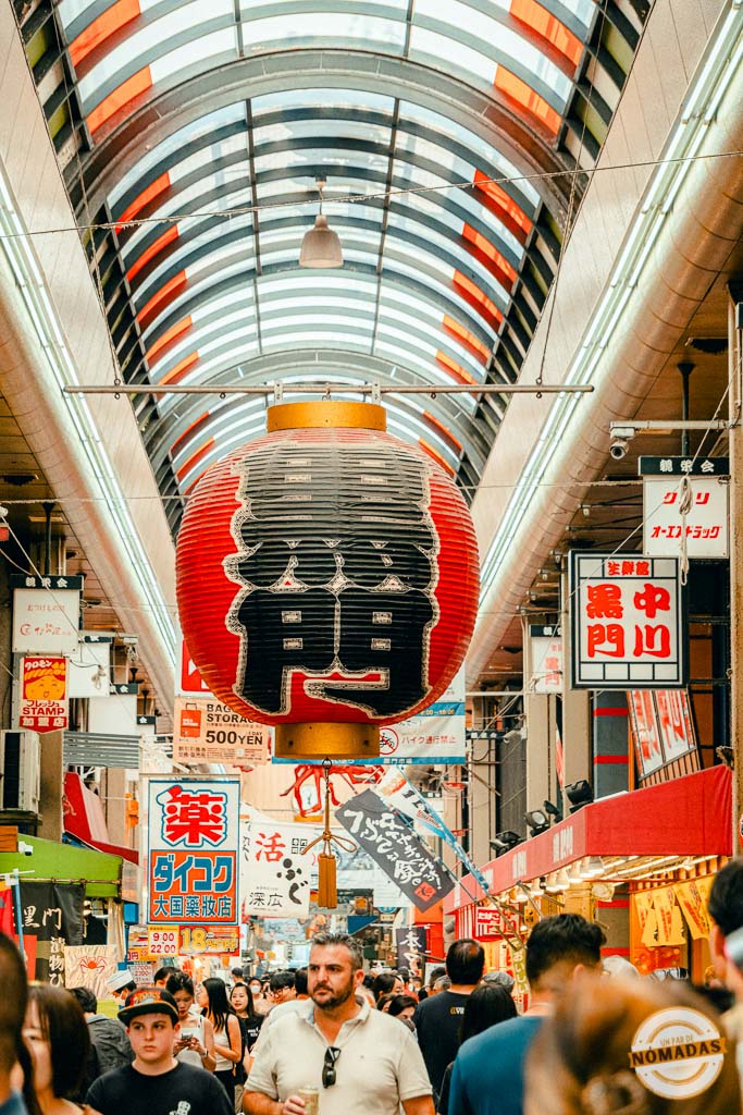 Gran farol rojo colgante en el mercado Kuromon de Osaka con una multitud de personas recorriendo los puestos bajo techo
