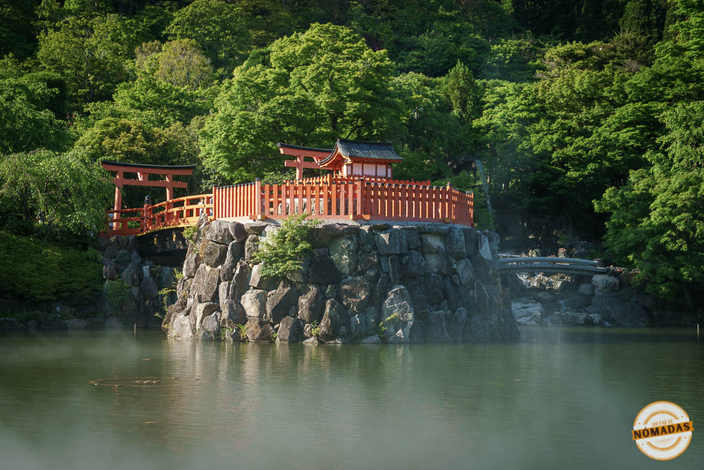 Santuario japonés rojo sobre una isla rocosa rodeado por lago y árboles verdes.
