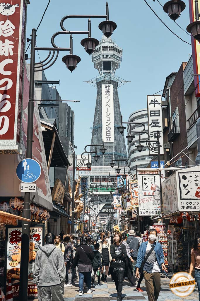 Vista diurna de la Torre Tsutenkaku en Osaka desde una calle comercial en Shinsekai, con gente paseando y letreros tradicionales
