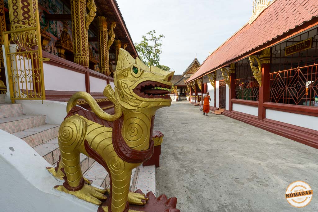 Estatua de león guardián en la entrada del Wat Sene, un templo budista en Luang Prabang, con un monje caminando al fondo.
