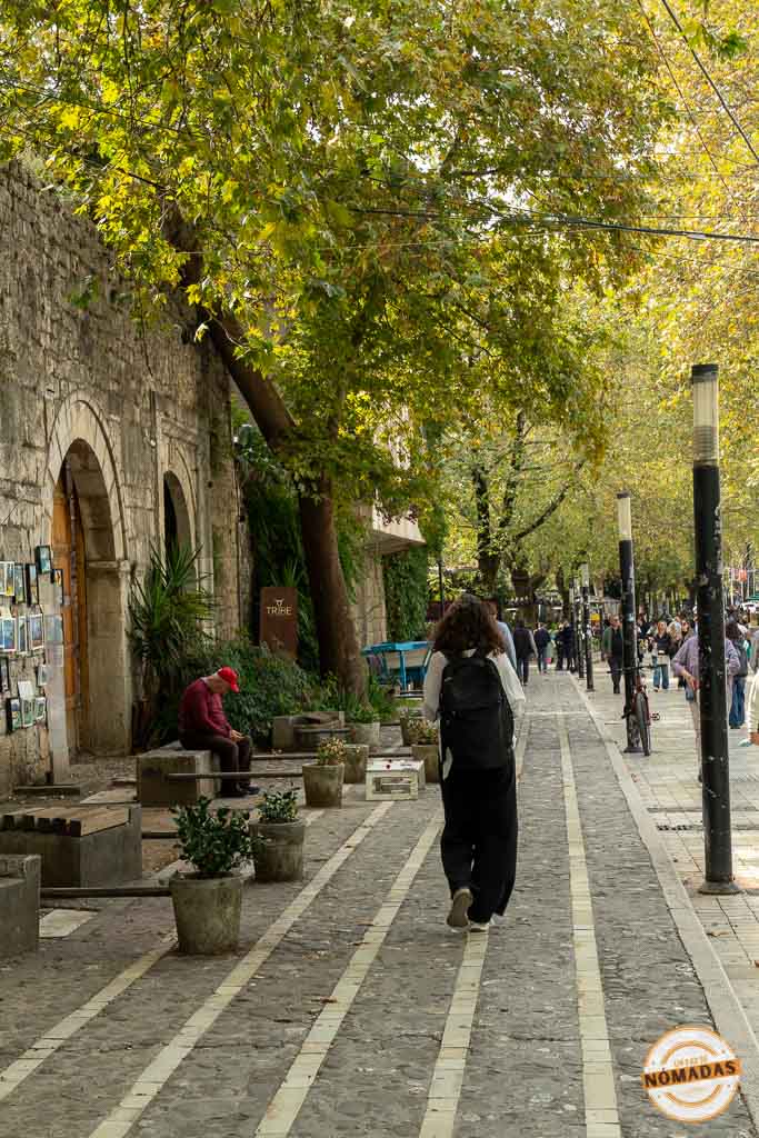 Gente paseando tranquilamente por la calle peatonal Murat Toptani en Tirana, bajo la sombra de los árboles y junto a muros de piedra históricos.