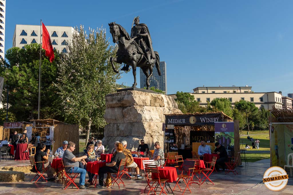 La estatua ecuestre de Skanderbeg en el centro de Tirana, rodeada de gente sentada en mesas rojas disfrutando de una feria de vino al aire libre.