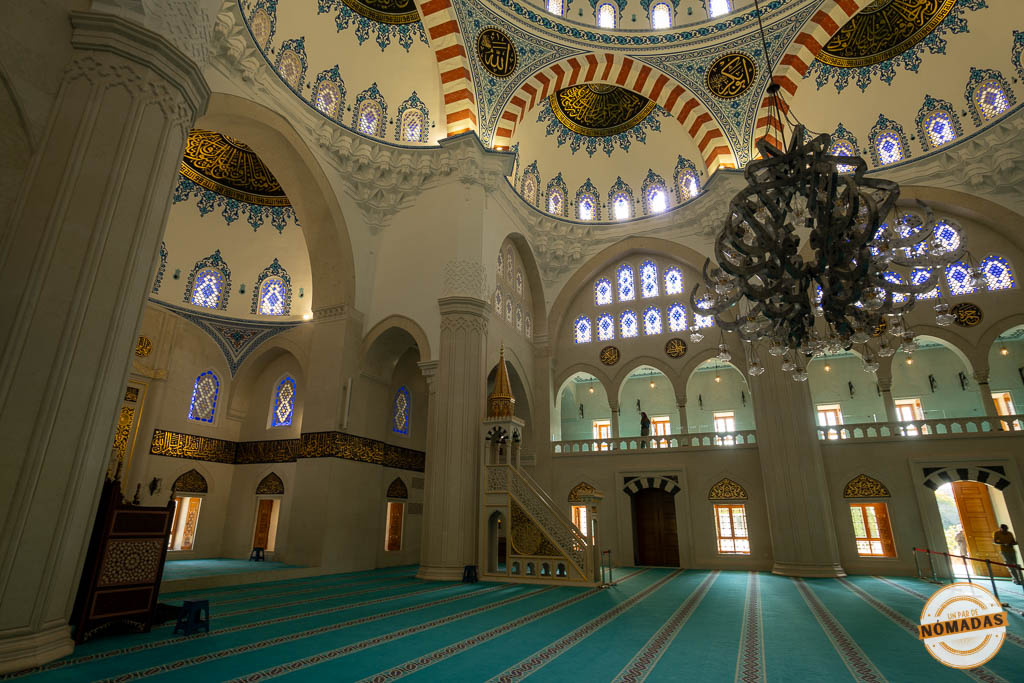 Vista interior de la sala de oración de la Gran Mezquita de Namazgah, con una enorme cúpula decorada, grandes lámparas colgantes y alfombra verde.