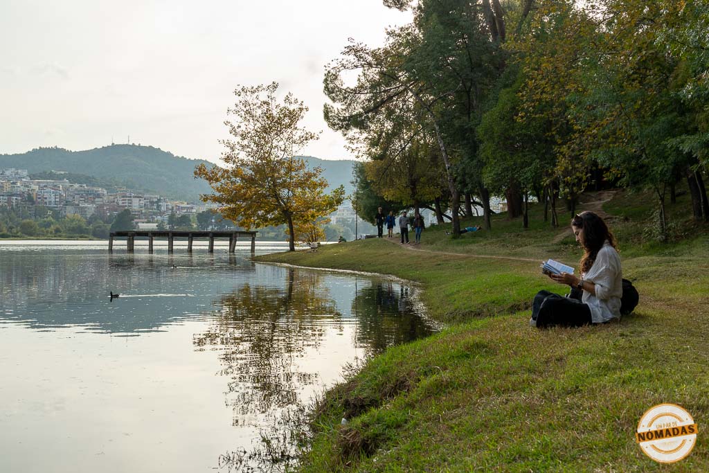 Tatiana leyendo tranquilamente sentada en el césped a orillas del Lago Artificial de Tirana, con un muelle de madera al fondo.