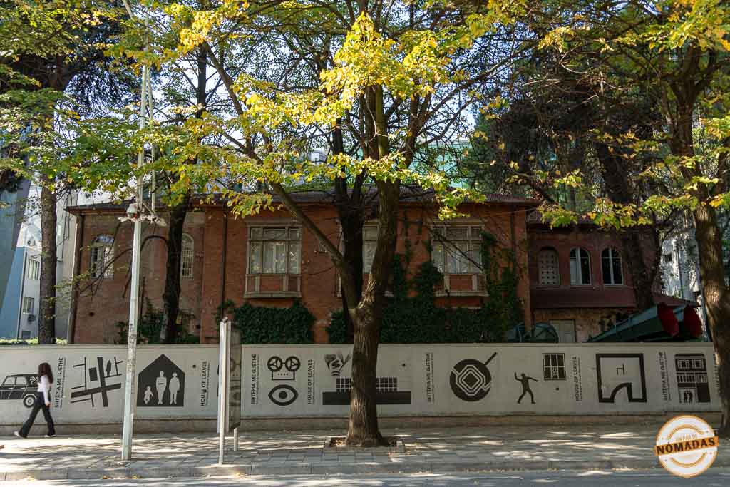 Fachada de ladrillo rojo del Museo de la Casa de las Hojas en Tirana, con árboles y un muro exterior decorado con iconos sobre el espionaje.