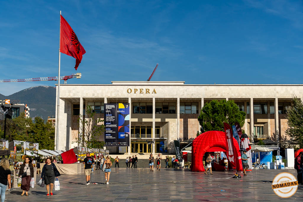 Fachada del Palacio de la Cultura y la Ópera en la Plaza Skanderbeg, con la bandera de Albania ondeando y ambiente de mercado.