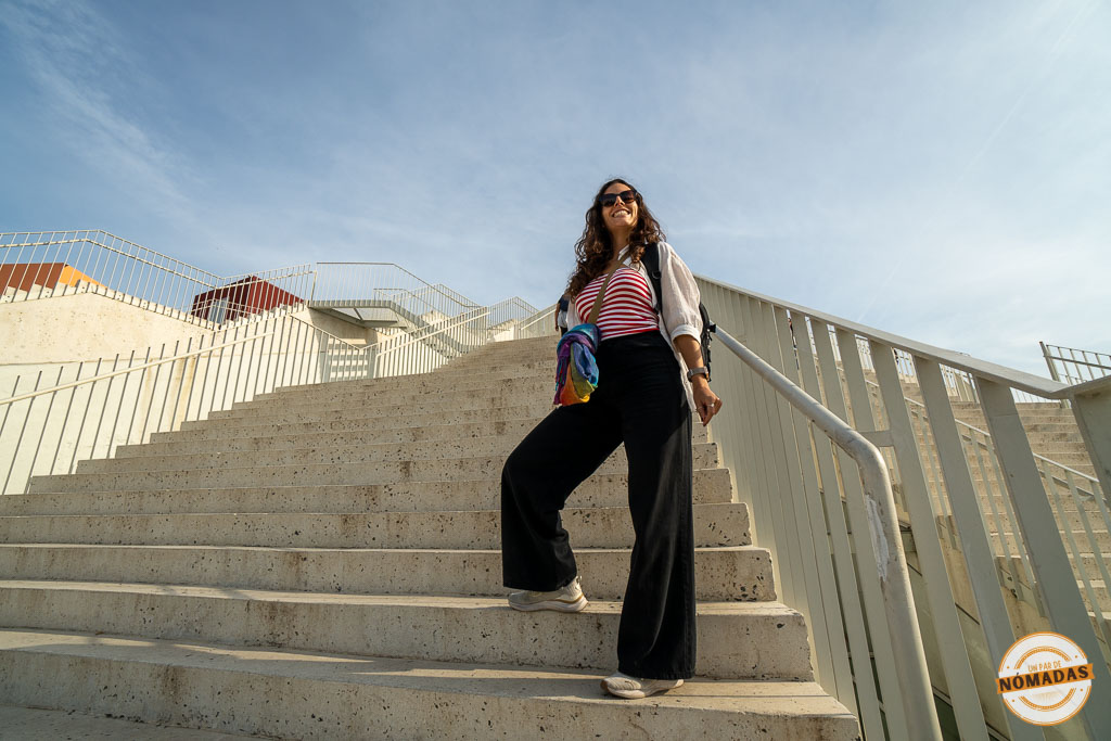 Tatiana subiendo las escaleras exteriores de la renovada Pirámide de Tirana, un edificio de hormigón blanco bajo un cielo azul.