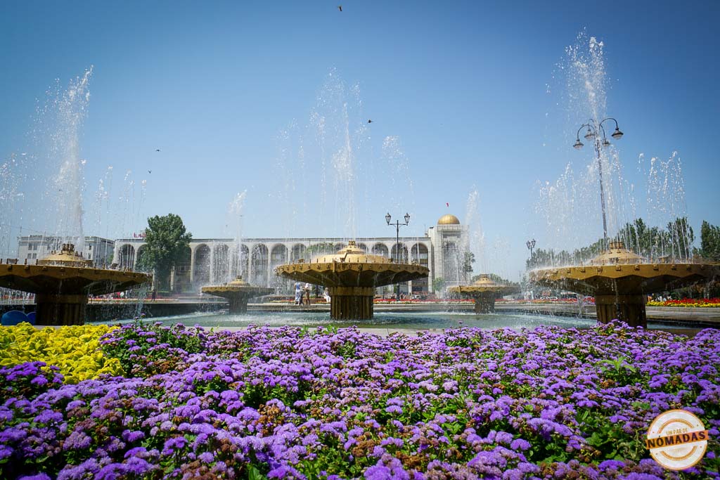 Primer plano detallado de flores moradas vibrantes en un parterre de la Plaza Ala-Too, con grandes fuentes de agua y un edificio monumental de fondo, perfecto para un resumen sobre qué ver en Bishkek.
