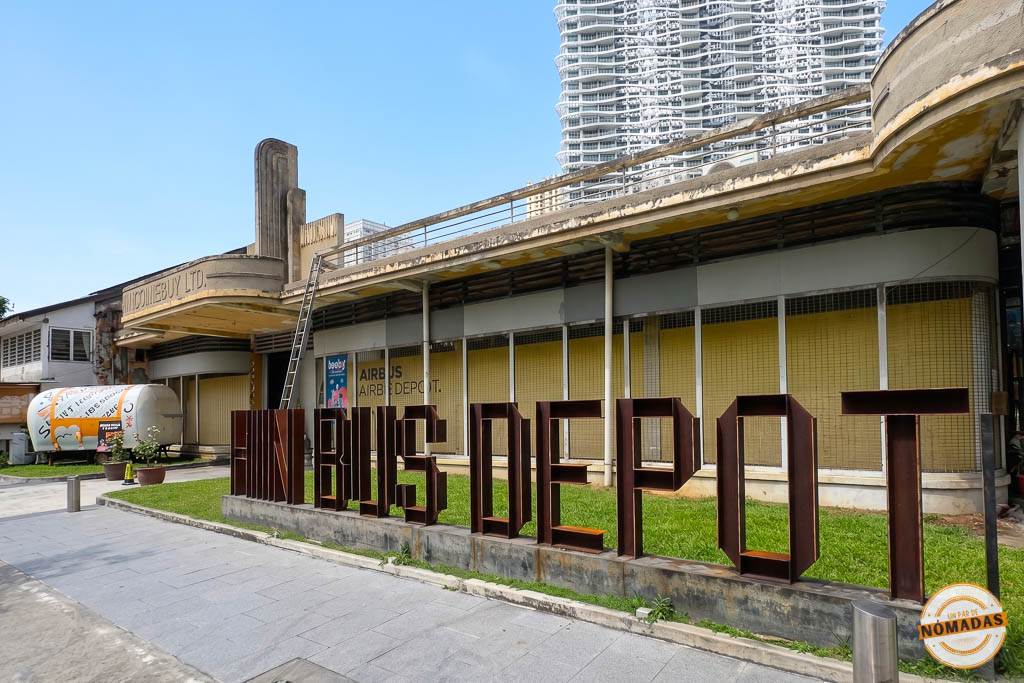 Grandes letras de metal oxidado que forman el nombre de Hin Bus Depot frente a un edificio art decó restaurado, con un moderno rascacielos asomando al fondo en Georgetown, Penang.