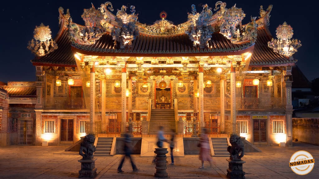 Fachada de la histórica casa de clan Khoo Kongsi en Georgetown profusamente iluminada por la noche, mostrando su intrincado tejado tallado, pilares y personas caminando con efecto de movimiento en el patio.