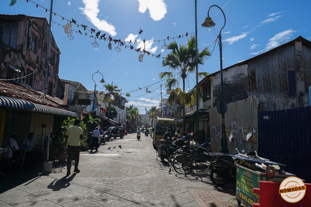 Vista de perspectiva de la soleada calle Love Lane en Georgetown, Penang. Muestra edificios antiguos desgastados, cables aéreos con decoraciones de siluetas de pájaros, palmeras, motos aparcadas y gente paseando bajo un cielo azul brillante.