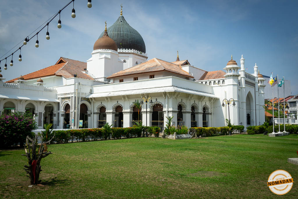 Fachada blanca con arcos y cúpulas oscuras de la Mezquita Kapitan Keling, rodeada de un gran jardín verde en el centro de Georgetown, Penang.