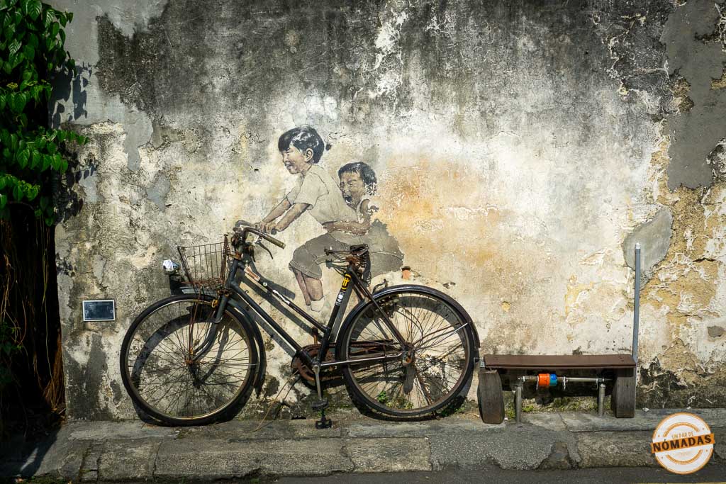 Mural interactivo de arte callejero del artista Ernest Zacharevic en Georgetown, Penang, que muestra a dos niños sonrientes pintados en una pared desgastada, que parecen estar montando una bicicleta negra física real apoyada contra la pared. Hay una pequeña banca con ruedas de colores a la derecha.