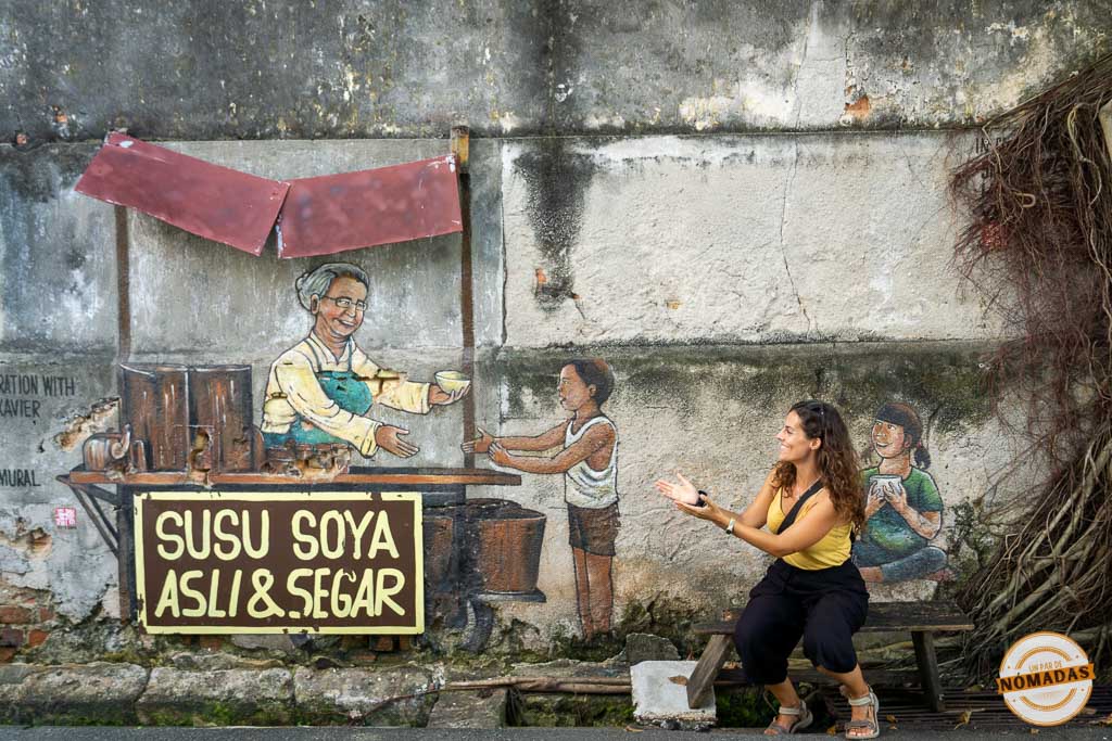 Viajera sonriente interactuando con un mural de arte callejero en Georgetown que representa un puesto tradicional de leche de soja (Susu Soya Asli & Segar). La viajera está sentada en un banco de madera físico frente a la pared desgastada, extendiendo las manos hacia los personajes pintados.