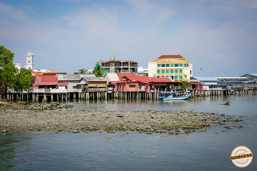 Casas tradicionales de madera y chapa construidas sobre pilotes de madera en el agua en los Clan Jetties de Georgetown, con edificios modernos y una torre del reloj al fondo.