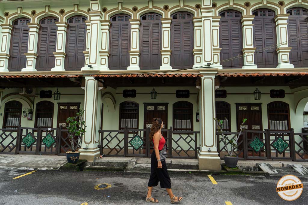 Viajera caminando por la calle frente a una elegante hilera de shophouses tradicionales de estilo Peranakan, pintadas de color verde pálido con grandes contraventanas de madera oscura en Georgetown, Penang.