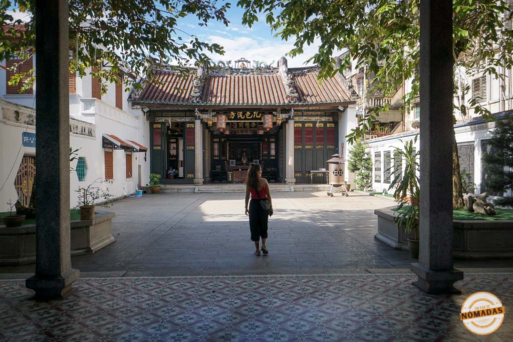 Viajera caminando por el soleado patio del Templo Ancestral de Han Jiang en Georgetown, Penang. La escena está enmarcada en primer plano por columnas de piedra y un suelo de baldosas con motivos geométricos.