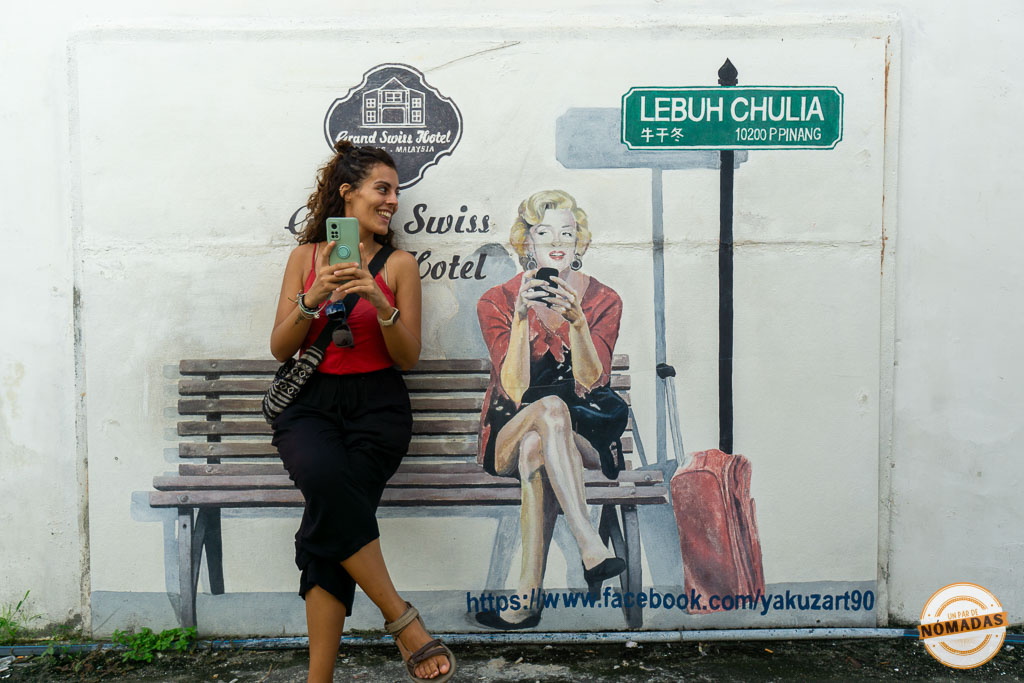 Viajera sonriente interactuando con un mural de arte callejero de 'yakuzart90' (Maldybayev) en Georgetown, Penang. El mural, en una pared blanca, muestra a una mujer pintada sentada en un banco de madera, ambas mirando sus smartphones. Incluye señalización de 'LEBUH CHULIA', el logotipo de 'Grand Swiss Hotel', una maleta pintada y un enlace de Facebook. La viajera física imita la pose y sonríe a la figura pintada. Esta escena interactiva en la calle Chulia es un ejemplo clave del arte callejero que ver en Penang, Malasia.
