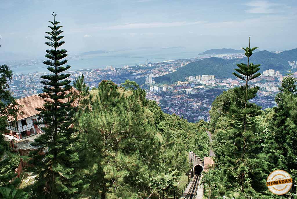 Vistas panorámicas de la ciudad de Georgetown y el mar desde la cima de Penang Hill, con las vías del histórico funicular y gran vegetación en primer plano.